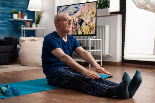 Senior man stretching legs muscle while sitting on yoga mat in living room during pilates workout. Pensioner training body flexibility slimming weight exercising gymnastic exercise