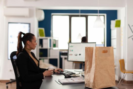 Close up shot of paper bag with tasty delicious meal takeaway food, of business woman working on computer analyzing company documents, graphs. Employee holding coffee cup.