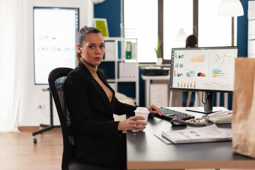 Portrait of business woman in startup company office, looking at camera holding cup of coffee. Paper bag from fast food with tasty food. Lunch packed in delivery bag.