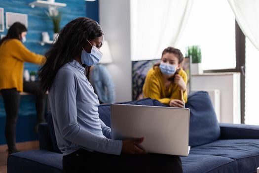 Cheerful happy african woman showing video on laptop to friend while hanging out in apartment living room keeping social distancing due to coronavirus pandemic to prevent virus spread.