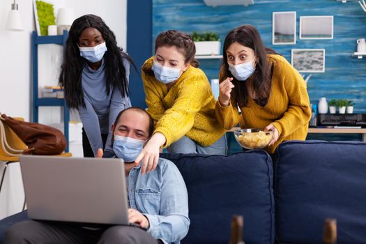 Group of mixed race friends having fun keeping social distancing with face mask preventing coronavirus spreading looking shocked at video on laptop in living room drinking beer and eating chips. Conceptual image.