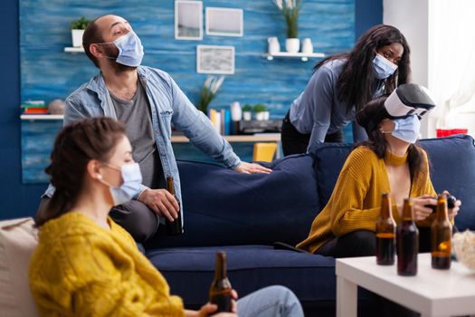 African woman cheering up for friend while playing video games wearing face mask during social pandemic to prevent illness with covid. Group of multiethnic people hanging out using vr headset.