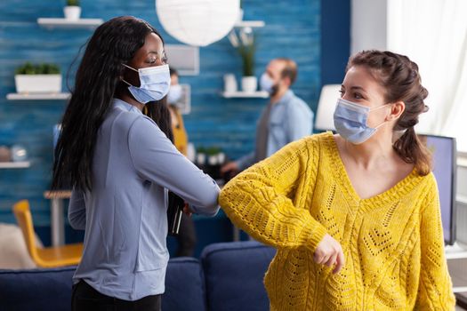 Cheerful african woman and her friend touching elbow keeping social distancing while greating each other wearing face mask to prevent coronavirus spread in the course of global pandemic in living room.