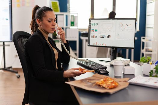 Multitasking businesswoman talking at landline explaining company graphs to remote collegue while having takeaway delivery meal food order at desk. Manager eating tasting pizza slice