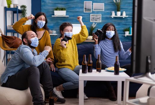 Multiethnic friends celebrating victory while playing video games wearing face mask and keeping social distancing during global pandemic with corona in home living room raising hands.