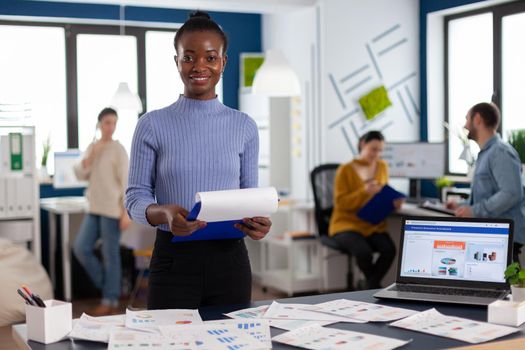 Smiling black woman entrepreneur holding clipboard with company strategy smiling at camera. Diverse team of business people analyzing company financial reports from computer.