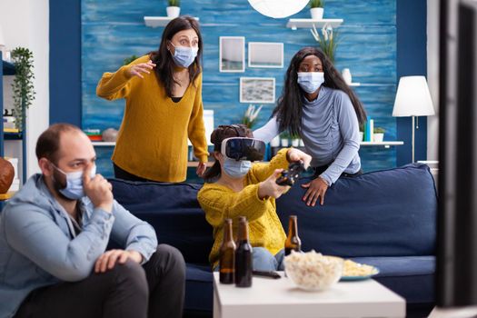 Multiethnic diverse friends enjoying vr tehnology playing video games in living room wearing face mask to prevent infection with coronavirus keeping social distancing. Conceptual image.