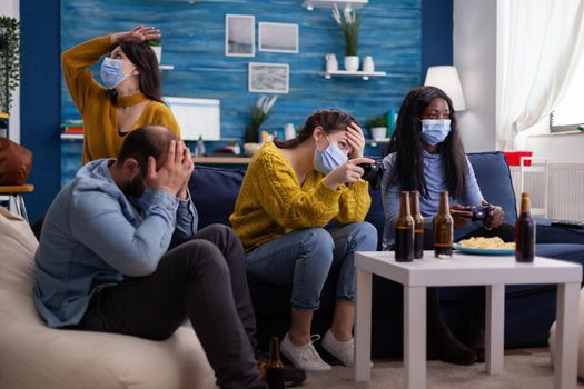 Disappointed diverse group of people playing video games respecting social distancing because of orona outbreak wearing face mask not to spread the virus and get sick. Beer bottle and chips.