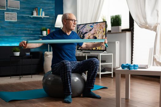 Invalid pensioner practicing active arm exercises using resistance elastic band watching online fitness workout video on laptop. Pensioner working body healthcare sitting on swiss ball in living room