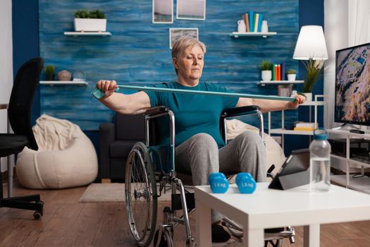 Invalid old woman in wheelchair stretching arm resistance training body muscle using elastic band after leg accident watching therapy video on tablet. Pensioner exercising gym workout in living room