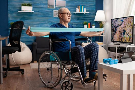 Disabled senior man in wheelchair training with elastic band exercising body workout recovering after disability accident watching strech video on tablet. Pensioner doing healthcare arm exercise