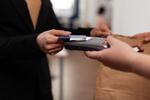 Close up of business woman holding credit card of pos, using contactless technology. Customer paying for their order with a credit card. Holding takeaway food in paper bag.