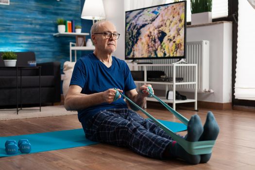 Retirement senior man sitting on yoga mat stretching legs muscles using resistance elastic band training body flexibility. Pensioner in sportswear slimming weight during muscle training in living room