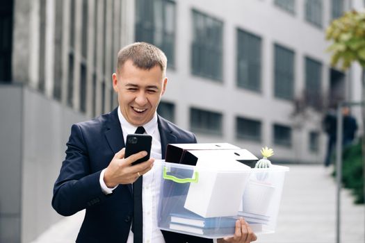 Businessman with box of personal stuff uses phone texting scrolling tapping. Happy businessman stand smiling use phone near business center. Portrait suit career male office handsome technology.