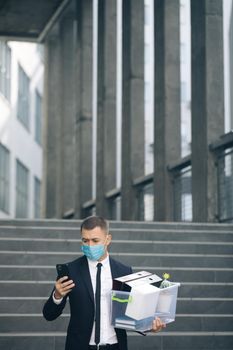 Businessman in medical mask with box of personal stuff walking the street use phone. Business style suit. Coronavirus outdoors social distancing. Finance and industry. Fired man lost job.