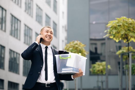 Happy man talking on phone with stuff in box. Cellphone conversation. Speaking on telephone. An employee between office buildings with a box and documents with a desk flower