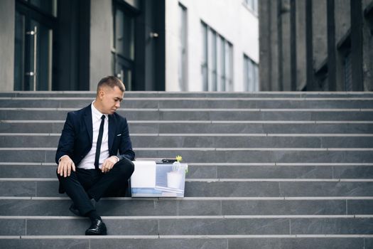 Male office worker in despair lost job. Sad businessman sitting on stairs outdoor with box of stuff as lost business. Fired man. Unemployment rate growing due pandemic.
