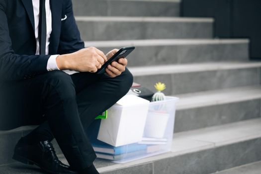 Sad businessman sitting on stairs outdoor with box of personal stuff. Despaired male lost work place and looking for a job on smartphone. Pandemic economical crisis. Fired man.