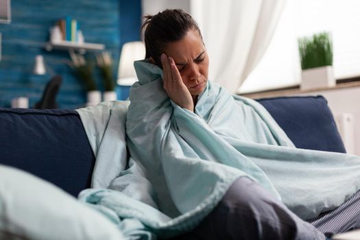 Caucasian woman feeling sick at home with headache, taking medical treatment. Adult in pijamas having cold flu illness sitting on sofa tired, with migraine and sore throat blowing nose