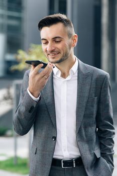 Businessman use smartphone to send voice messages outdoors at downtown. Young business man talking on phone near modern office building. Vertical screen orientation