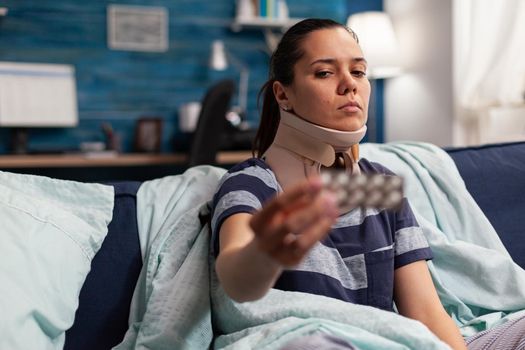 Woman with sickness and cervical foam collar on couch taking medical treatment for back and neck pain. Caucasian adult with muscular contracture after physical injury in accident