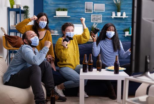 Excited group of multiethnic friend celebrating victory while playing video games in home living room keeping social distancing wearing face mask not to spread corona in time of global pandemic.
