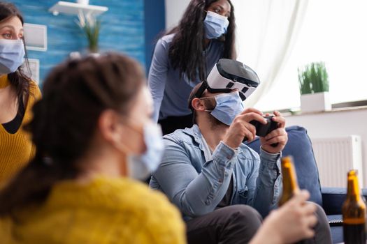 Excited man spending time with friends experiencing vritual reality playing games with vr headset wearing face mask to prevent coronavirus spread in time of social pandemic.