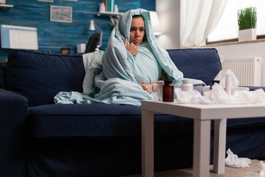 Suffering woman in blanket feeling ill at home with seasonal virus infection symptoms. Unwell caucasian young person resting on couch with sickness disease headache wrapped in blanket