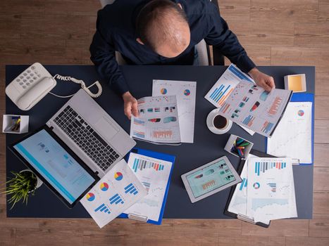 Top view of entrepreneur man holding and analyzing financial paper sitting at desk in business office thinking business strategy. Laptop with finance statistics.