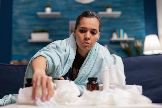 Sick woman taking medicine for seasonal virus wrapped in blanket holding pills. Caucasian young person treating disease with medical treatment for covid 19 symptoms temperature pain