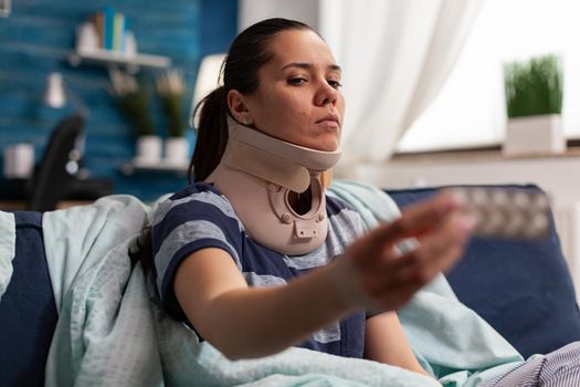 Woman with sickness and cervical foam collar on couch taking medical treatment for back and neck pain. Caucasian adult with muscular contracture after physical injury in accident