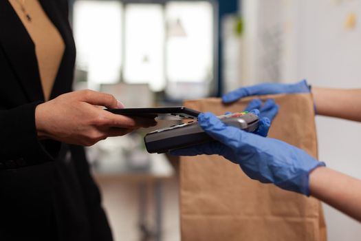 Delivery guy wearing protective gloves receiving paymant from businesswoman using smartphone nfc, technology over pos terminal service for delicious tasty meal lunch in paper bag.