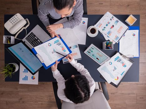 Top view of manager explaing financial strategy to employee sitting at desk in corproate office, pointing at charts on clipboard. Coworkers during briefing in start up company.