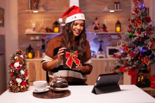 Joyful woman holding present box while on video call conference concept with friends in festive kitchen. Caucasian young adult receiving surprise gift for christmas eve seasonal festivity