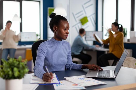 African businesswoman taking notes on clipboard analysing statistics in start up office. Diverse team of business people analyzing company financial reports from computer. African businesswoman taking notes on clipboard analysing statistics