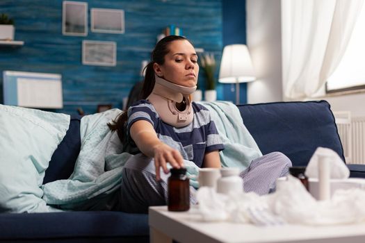 Woman with cervical neck collar sitting on sofa suffering from spine pain. Young caucasian person with physical injury taking pain medicine after back accident, with discomfort