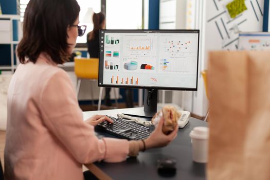 Businesswoman eating tasty sandwich having meal break working in business company office during takeout lunchbreak. Fast food order paper bag delivered at workplace. Woman typing strategy on computer.