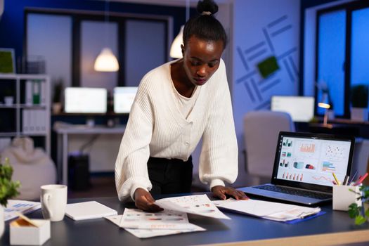 Stressed african manager woman working with financial documents standing at desk checking graphs, holding papers, reading raports late at night in start-up office doing overtime to respect deadline.