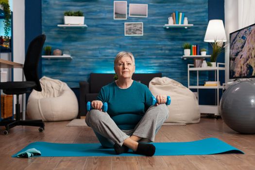 Portrait of athlete senior woman looking into camera sitting in lotus position on yoga mat in living room during wellness workout. Retired pensioner training body muscles holding dumbbells