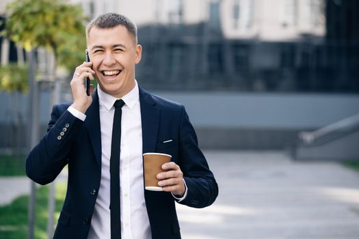 European businessman talking on mobile phone walking near office building background. Man is in dialogue, smiling, holding coffee cup on summer day.