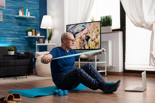 Pensioner working at body resistance exercising arms muscles using elastic band sitting on yoga mat with crossed legs position. Senior man doing workout during fitness class looking on laptop