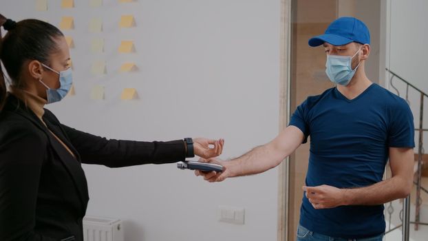 Delivery guy with protection face mask and gloves delivering takeaway food box during lunchtime in company office. Businesswoman client paying meal order with smart watch using POS contactless