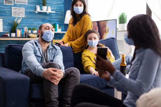 African woman showing funny clip on smartphone hanging out in home living room wearing face mask preventing coronavirus during global pandemic keeping social distancing.