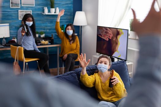 Multiethnic group of people greeting friend waving at him keeping social distancing wearing face mask to prevent infection with covid19, holding beer bettle in living room. Conceptual image.