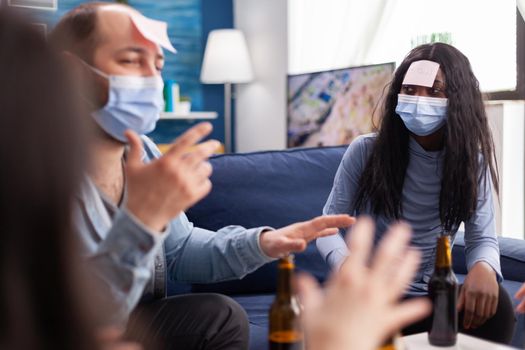 African woman with sticky note on forehead playing name game with cheerful happy friends wearing face mask to prevent infection with covid19 due to social pandemic. Beer bottle,