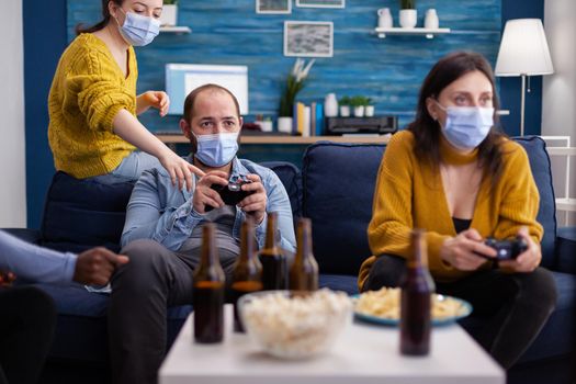 Diverse friends trying to win playing video games using joystick havin fun wearing face mask to prevent spreading of coronavirus in time of global outbreak. Gaming competition, beer and popcorn.