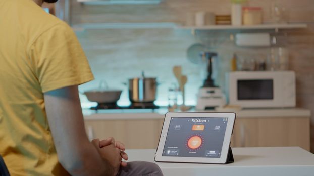Man looking at tablet with intelligent software placed on kitchen table controlling light with high tech application. Person using notepad with smart home app in house automation system