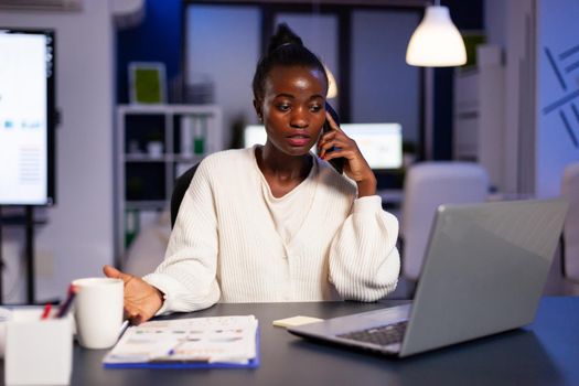 African employee speaking at phone while working at laptop late at night. Busy focused freelancer doing overtime for job reading writing, searching, respecting deadline of financial project, overworking.