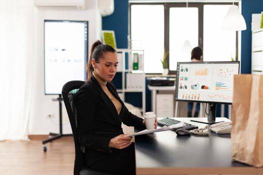 Businesswoman in corporate start up office analyziing documents, chart looking at clipboard. Recycled paper bag with delicious tasty food for lunch break. Employee holding coffee cup.