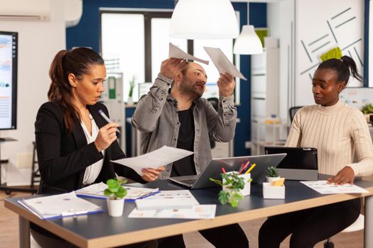 Angry business man screaming at diverse colleagues during office meeting throwing away documents, disagreeing about bad business contract. Young multiracial group of people working in coworking space.